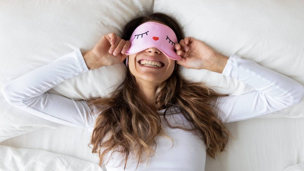 Top view close up happy woman wearing funny sleeping mask enjoying morning in bedroom, lying in comfortable bed on soft pillow, beautiful girl with healthy toothy smile stretching in the morning