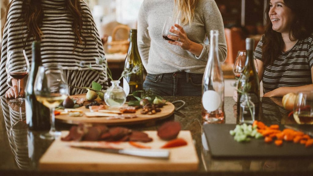 Two women standing beside a woman sitting at a table with food.