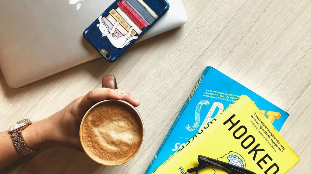 Hand holding coffee with laptop, phone, books and glasses on the table.