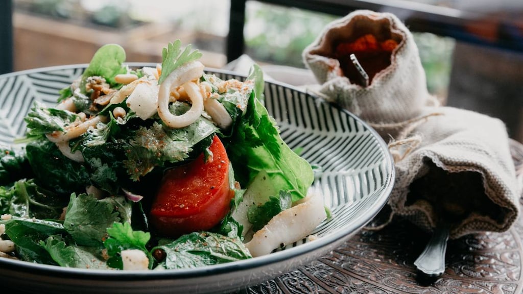 A bowl of salad and seafood sitting on a table with cutlery wrapped in a napkin.
